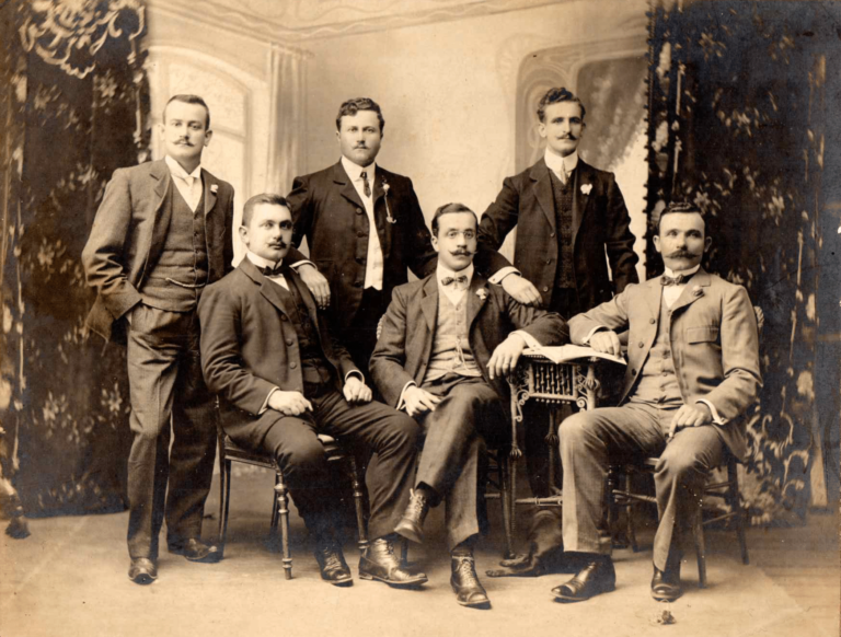 Retrato de grupo de siete hombres con traje, en un interior con cortinados, a comienzos del siglo XX